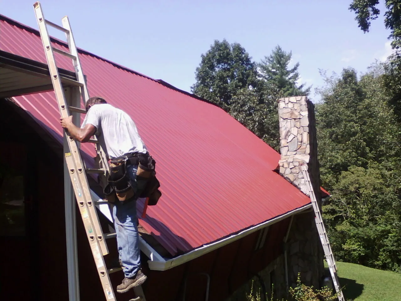 Roofer on a ladder beside a red metal roof and stone chimney — Soffit & Fascia Repair by Roofing Brothers in Sparrow Bush